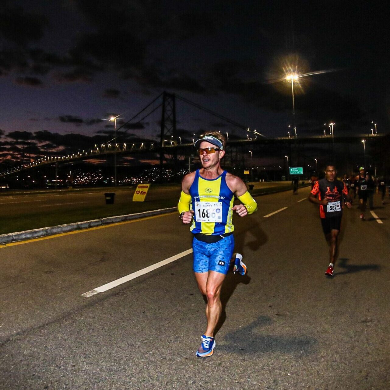 A runner in a blue and yellow outfit (Coach Peru) competes in a night marathon, with a bridge and city lights in the background.
